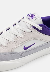 Bijele i sive tenisice s ljubičastim akcentima, s gornjim dijelom od antilopa i platna, vezicama i vidljivim Nike logotipom na jeziku.