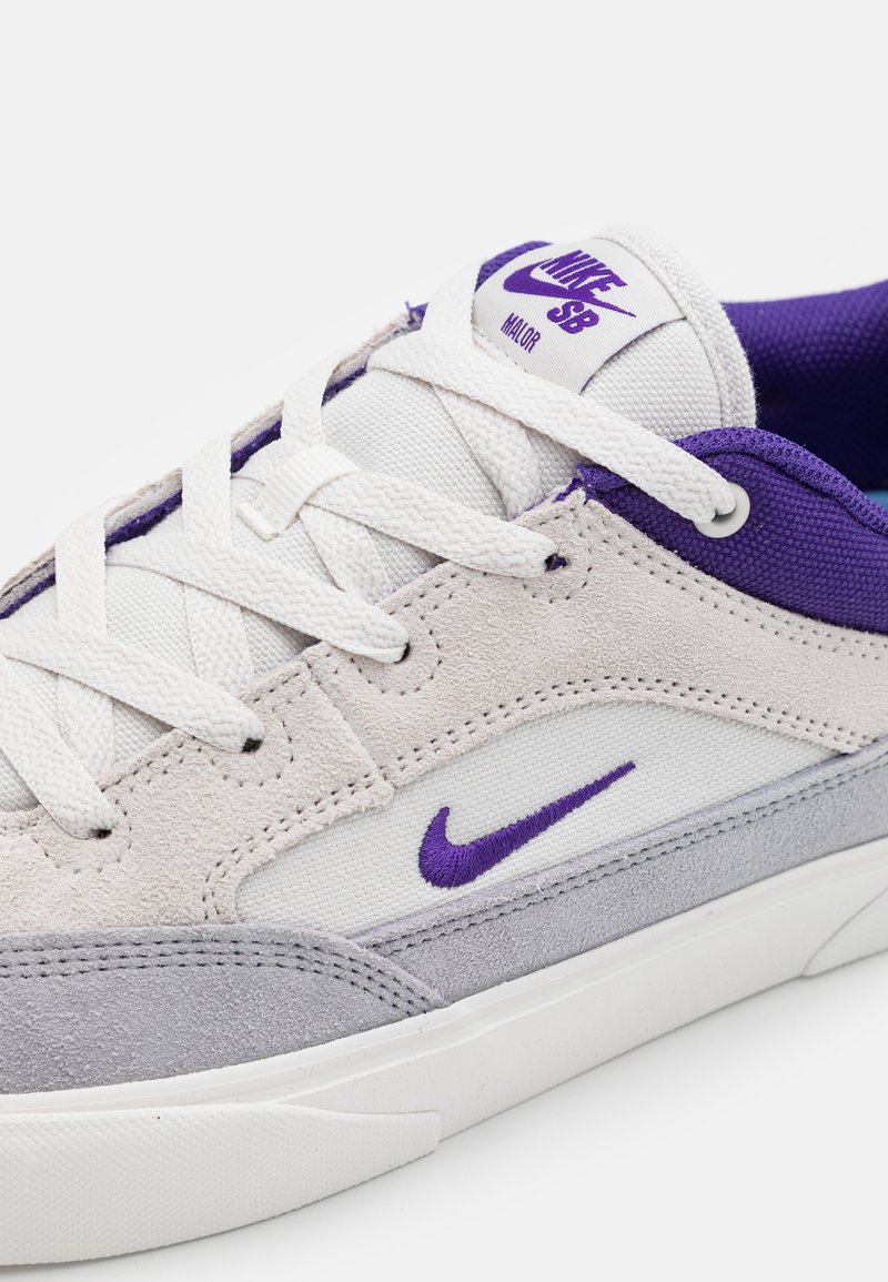 Weißes und graues Sneaker-Modell mit lila Akzenten, das ein Obermaterial aus Wildleder und Canvas, Schnürsenkel und ein sichtbares Nike-Logo auf der Zunge aufweist.