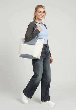 Joven sonriente, vestida con una camiseta de manga larga azul y gris, vaqueros oscuros de pierna ancha, zapatillas blancas, llevando un gran bolso tote blanco con detalles azules.