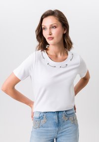 White short-sleeve t-shirt featuring a round neckline adorned with silver hardware accents. Smooth texture with a casual fit.
