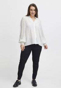 Weiße, langärmlige Bluse mit Knopfleiste, dezenten vertikalen Streifen und einem besetzten Spitzenkragen, kombiniert mit schwarzen Slim-Fit-Hosen und schwarzen Schuhen.