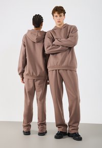 Brązowa bluza z kapturem i dopasowane spodnie dresowe wykonane z miękkiego, teksturowanego materiału. Zakończone ściągaczami przy mankietach i luźnym kroju. Na zdjęciu czarne obuwie.