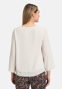 Blusa bianca con scollo rotondo e maniche a campana, caratterizzata da un orlo inferiore trasparente, abbinata a pantaloni marroni stampati. Texture liscia.