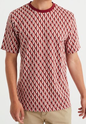 Mann trägt ein kurzärmeliges Hemd mit einem sich wiederholenden geometrischen Muster in Rot, Weiß und Pink, kombiniert mit beigen Hosen.