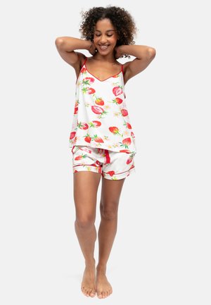 Jonge vrouw lacht met krullend haar, draagt een wit hemdje en korte broek met rood aardbeienpatroon, staat blootsvoets met handen achter het hoofd.