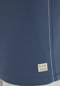 Kék pamut póló fehér varrással, amelyen egy anyagcímke található, amely a következő feliratot tartalmazza: "BLEND 1993 DENIM WITH ATTITUDE" a szegély közelében.