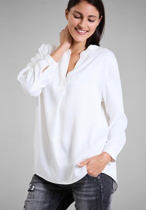 White blouse in smooth fabric, featuring a partial button placket and long sleeves. Worn with distressed gray jeans.