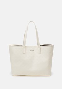 Creme tote taske med præget logo-mønster, glat tekstur og matchende håndtag. Har et sølvlogo på fronten. Ingen synlig hardware.
