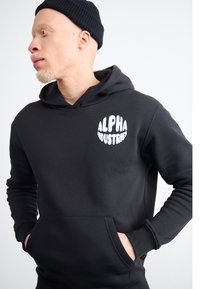 Hoodie preto com um bolso à frente, apresentando um logotipo circular branco "ALPHA INDUSTRIES". Tecido suave, corte padrão, com capuz ajustável por cordão.