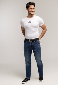 Młody mężczyzna uśmiechający się, ubrany w białą markową koszulkę, niebieskie dżinsy z czarnym paskiem oraz czarne trampki, stojący z rękami za plecami.