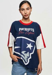 T-paita tummansinisellä pohjalla, punaisilla yksityiskohdilla, jossa on suuri Patriots-logo ja teksti "PATRIOTS New England" valkoisella. Vapaa-ajan malli, puuvillamateriaali.