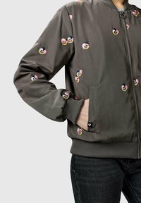 Grå bomberjakke med broderede blomster i pink, gul og sort, båret med hånden i lommen, kombineret med mørke jeans.