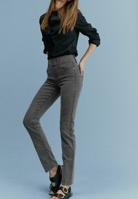 Frau in schwarzem, geknöpftem Shirt, grauen, figurbetonten Jeans und schwarzen Sandalen mit goldenen Schnallen, die mit der Hand in der Gesäßtasche vor einem blauen Hintergrund posiert.