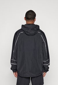 Mężczyzna ubrany w luźną czarną kurtkę z kapturem, z białymi detalami na rękawach, zwrócony tyłem na jednolitym jasnym tle.