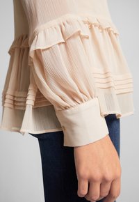 Manche de blouse beige transparente avec volants et plis, resserrée au large poignet, portée avec un jean bleu foncé sur fond gris.