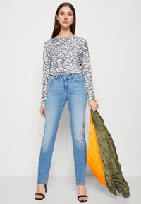 Top de manga larga con estampado de leopardo gris y blanco, jeans ajustados de color azul claro, sandalias negras de tacón, sosteniendo una chaqueta verde oliva y naranja.