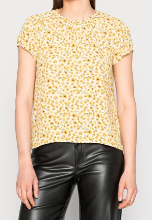 Mujer con blusa amarilla de flores y mangas cortas combinada con pantalones de cuero negro sobre un fondo liso.