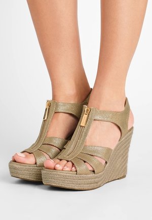 Pies con sandalias de cuña de textura dorada, con cremalleras frontales y suelas de plataforma trenzadas, sobre un fondo blanco.