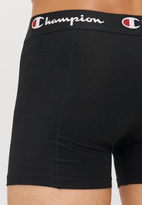 Black boxer shorts made of smooth fabric, featuring a wide waistband with "Champion" logo in white and red.