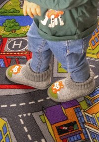 Un tout-petit portant des chaussons gris en forme d'animaux et un jean se tient debout sur un tapis de jeu coloré avec des routes et des bâtiments.