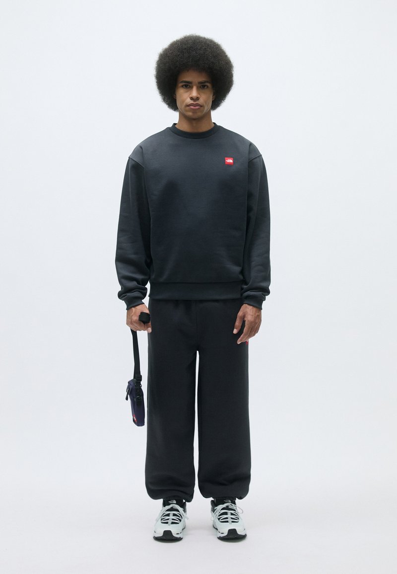 Mørkegrå sweatshirt- og buksesæt, lavet af blødt materiale, med et rødt logo-patch på brystet. Sneakers er hvide med sorte accenter.