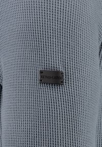 Grauer strukturierter Strickpullover mit einem rechteckigen schwarzen Etikett, auf dem der Markenname "KEY-LARGO" auf der Vorderseite angebracht ist.
