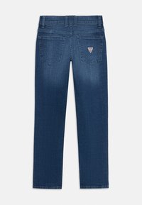 Jean en denim bleu foncé avec une coupe droite, comportant deux poches arrière et un petit patch logo au-dessus de la poche droite. Texture lisse.