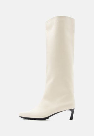 Λευκό μπότα μέχρι το γόνατο από λείο υλικό με μυτερή μύτη και λεπτό μαύρο τακούνι. Διαθέτει καθαρό σχεδιασμό και ελάχιστη σήμανση.