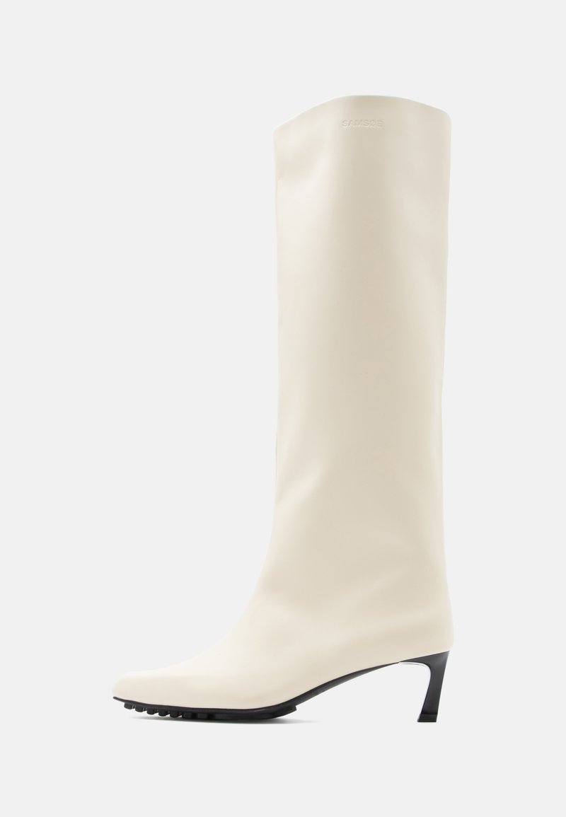 Λευκό μπότα μέχρι το γόνατο από λείο υλικό με μυτερή μύτη και λεπτό μαύρο τακούνι. Διαθέτει καθαρό σχεδιασμό και ελάχιστη σήμανση.