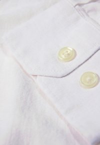 Witte linnen shirtstof met een puntkraag en twee doorzichtige knopen. De textuur lijkt licht gekreukt.