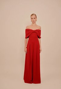Red off-the-shoulder jumpsuit features a bow detail at the chest, flowing fabric, and wide-leg design. Smooth texture with pleats.