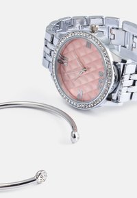 Sølvfarget klokke med en rosa teksturert urskive, romerske tall og rhinstein-detaljer; kombinert med et enkelt sølv armbånd.
