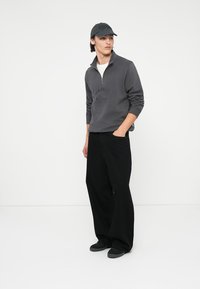 Sweatshirt cinza com zíper de um quarto e texto bordado, combinado com calças pretas largas e sapatos pretos, finalizado com um boné de baseball correspondente.