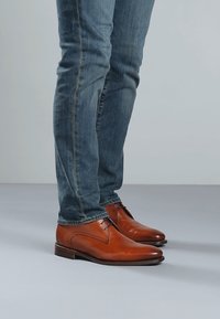 Person iført blå jeans og polerede brune lædersko, stående på en lys grå gulv mod en neutral baggrund.