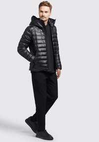 Geacă puffer neagră cu glugă, cu un design matlasat, textură lucioasă, închidere cu fermoar și buzunare laterale. Asortată cu pantaloni negri și adidași.