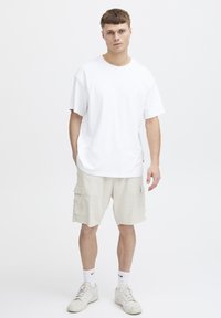Jonge man staat rechtop en kijkt naar voren, gekleed in een effen wit T-shirt, beige cargoshorts, witte Nike-sokken en witte sneakers voor een witte achtergrond.