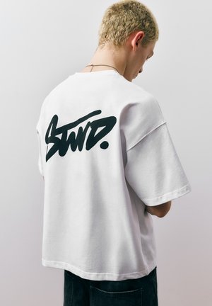 Mlada oseba s kratkimi blond lasmi, oblečena v belo preveliko majico z velikimi črnimi grafitnimi napisi na hrbtni strani.