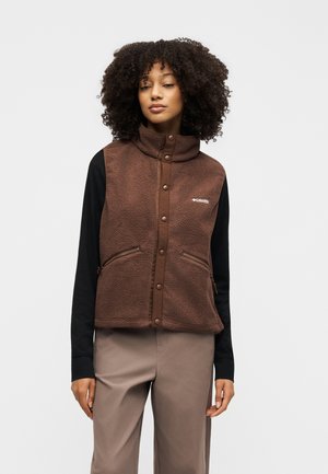 Femme aux cheveux bouclés portant un gilet en polaire marron par-dessus une chemise noire à manches longues et un pantalon beige, debout devant un fond uni.