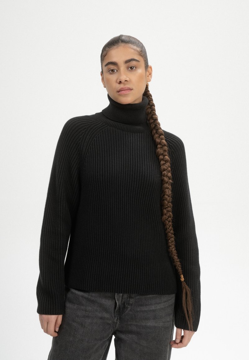 Schwarzer gerippter Strickpullover mit Rollkragen, langen Ärmeln und lockerer Passform, kombiniert mit dunklen Jeans. Verfügt über einen strukturierten Stoff.