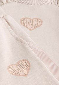 Világos bézs szövet puha textúrával, két szív alakú emblémával. Mindkét embléma finoman dombornyomott mintával az "ALLSAINTS" feliratot tartalmazza.