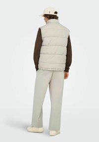 Lysegrå puffervest med høj krave, båret over en brun, struktureret striktrøje. Lyse bukser og en cremefarvet kasket afslutter outfittet.