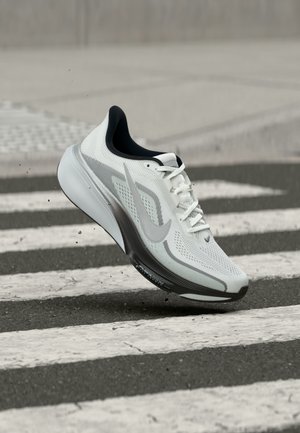 Chaussure de running Nike blanche et grise avec lacets en l'air au-dessus d'un passage piéton sur l'asphalte, affichant un mouvement dynamique et des détails de design.