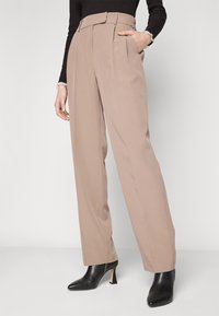 Pantalons beiges taille haute avec des plis, jambes larges et une coupe ajustée. Le tissu est lisse avec une légère brillance ; associés à des bottes à talons noires.