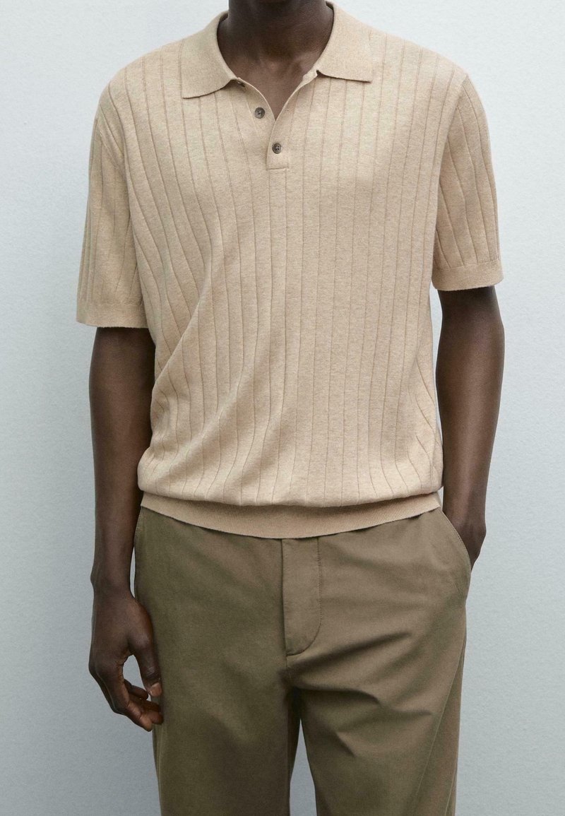 Homme portant un polo beige côtelé à manches courtes, rentré dans un pantalon vert olive, debout contre un mur de couleur claire.