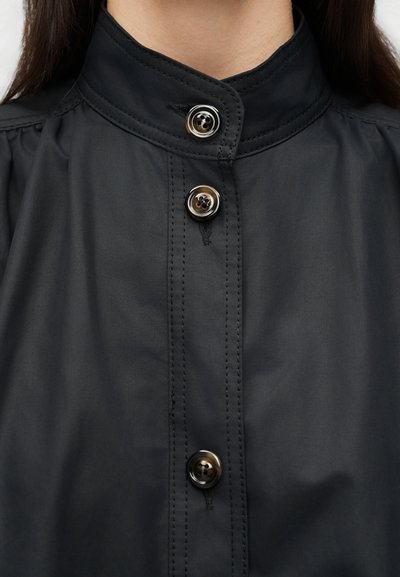 Gros plan sur le col d'une chemise noire à boutons, avec trois boutons nacrés et des coutures visibles sur un tissu lisse.