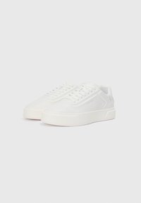 Adidași albi cu un superior din piele netedă, vârf rotund, șireturi plate și o talpă groasă din cauciuc. Branding subtil pe laterale.
