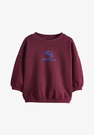 Jong kind met vlechten, draagt een bordeauxrode trui met een croissantlogo en zwart-wit geruite broek, staand tegen een lichte achtergrond.