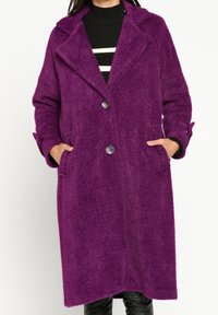 Lila, texturerad ullkappa med notched krage, två stora knappar och sidfickor. Har långa ärmar och en asymmetrisk fåll.