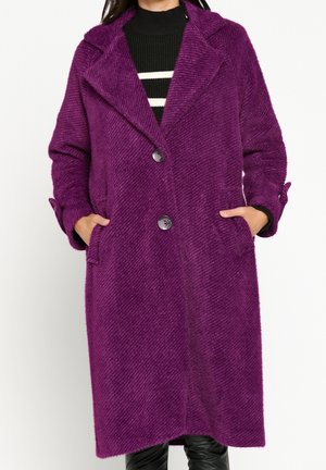 Lila, texturerad ullkappa med notched krage, två stora knappar och sidfickor. Har långa ärmar och en asymmetrisk fåll.