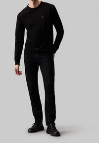 Zwart T-shirt met lange mouwen en een ronde hals, met een klein logo op de borst, gecombineerd met donkerblauwe jeans en zwarte veterschoenen.